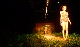 Model under a spotlight at the end of a fashion runway lined with foliage