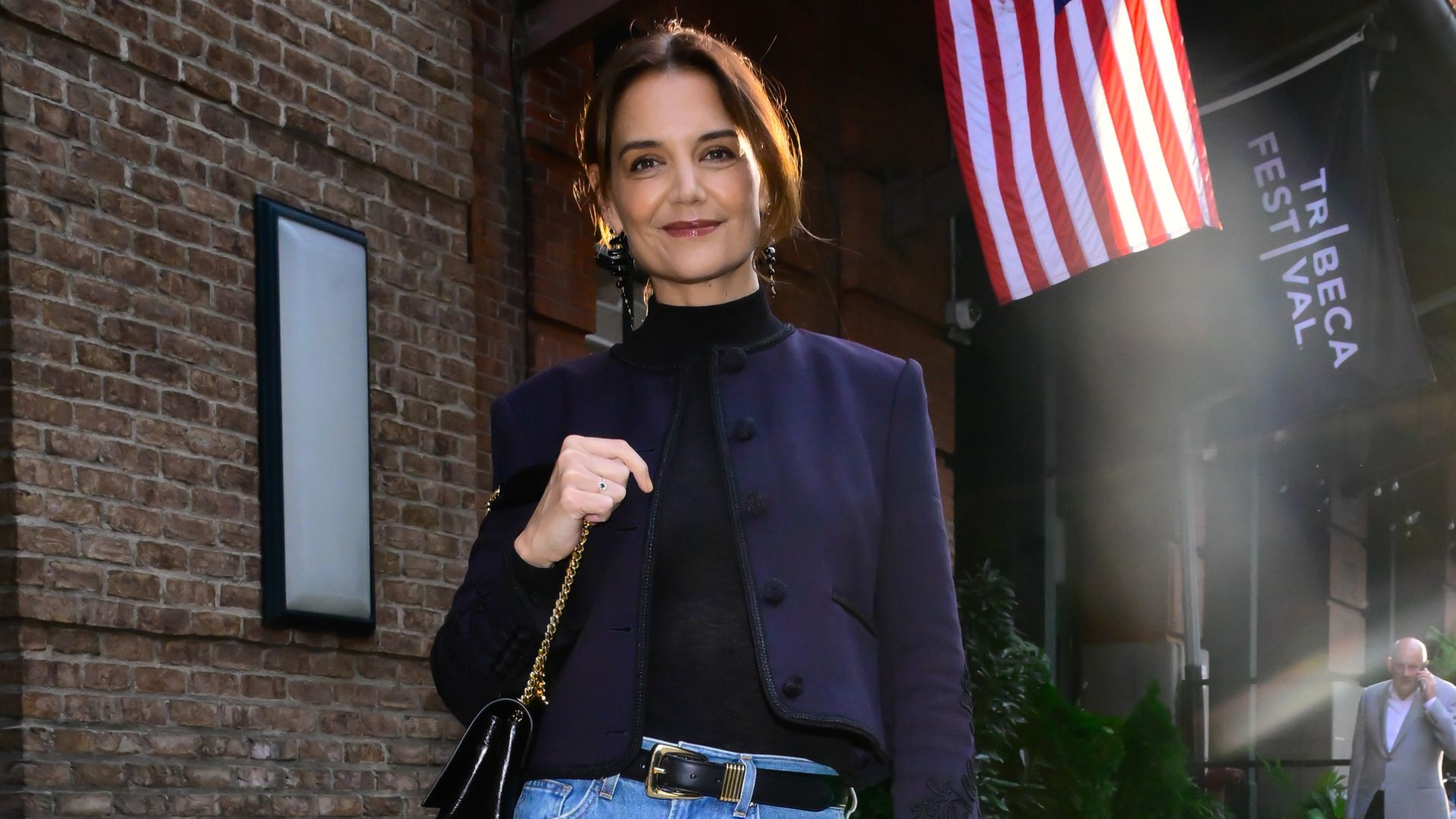 Katie Holmes stands outside on the pavement wearing blue jeans with black suede boots and a navy jacket.