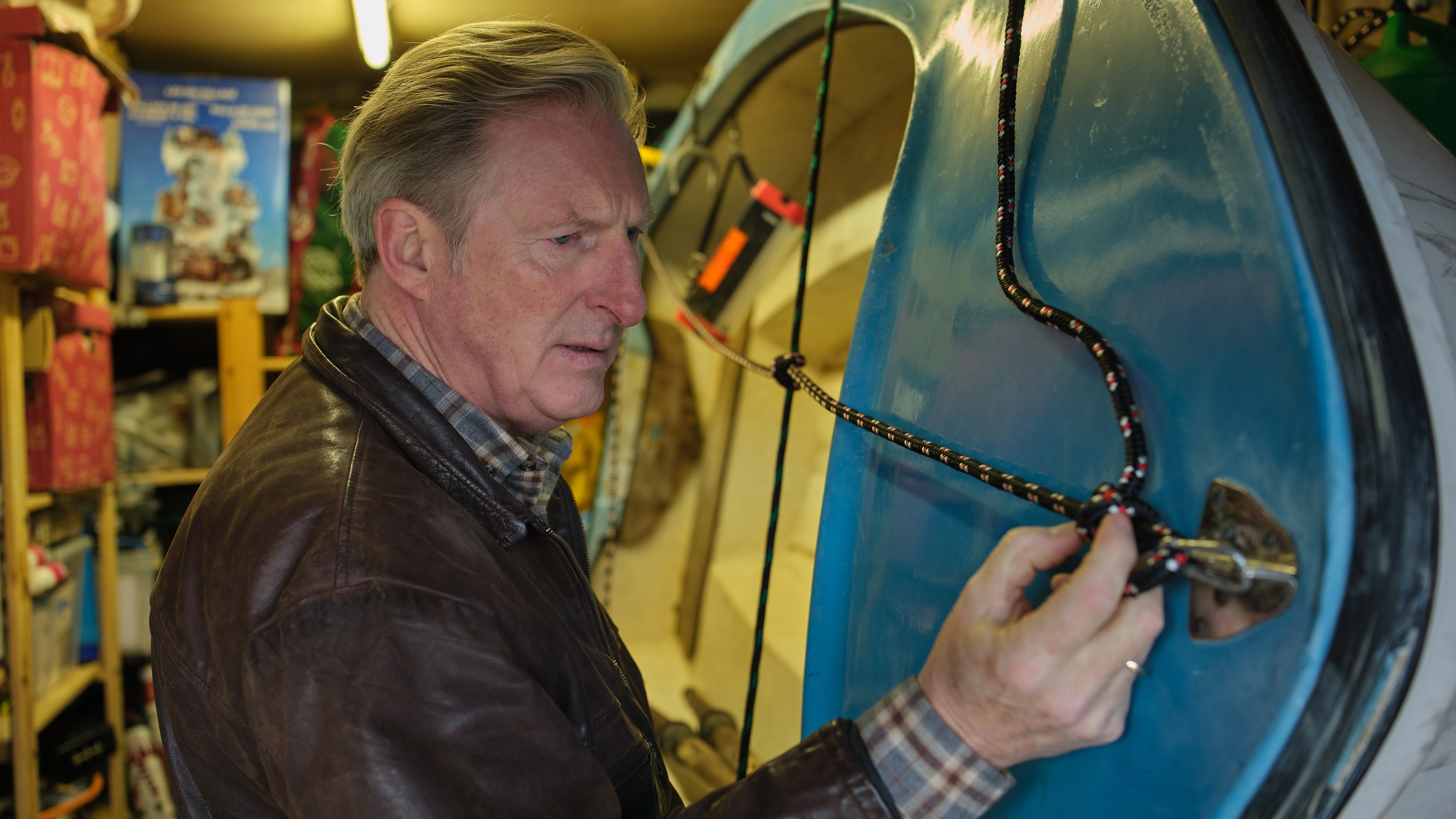 ADRIAN DUNBAR as Ridley looking at a knot on a rope around a boat and looking confused.