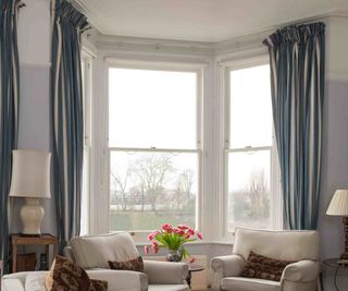 A period-style living room with a bay window with three sash windows by The Sash Window Workshop. The room features picture rails, blue and white curtains, comfy chairs and traditional-style furniture.