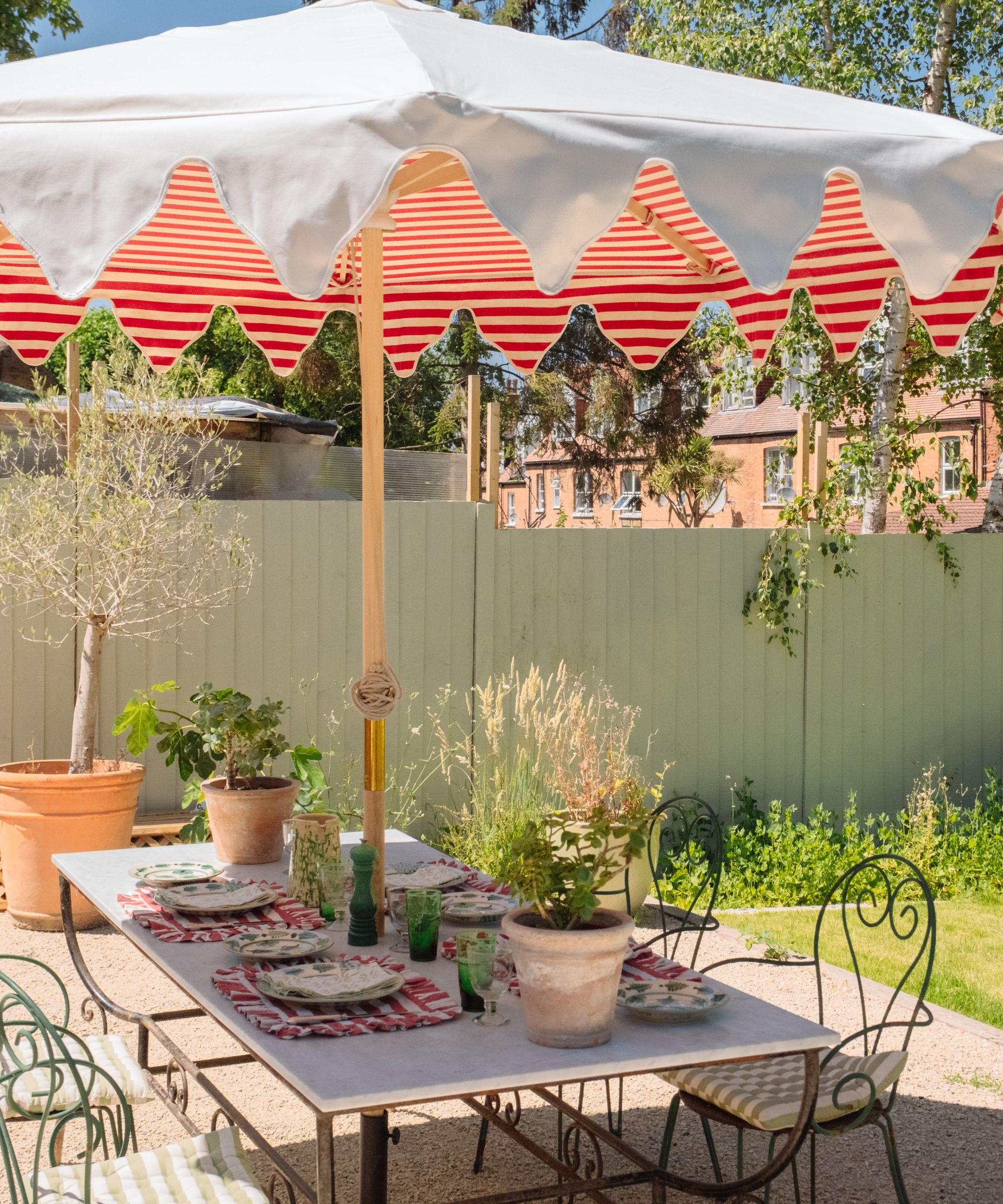 Garden with a sage green painted fence, marble and wrought iron long table decorated with plates, striped placemats and green glasses, wrought iron chairs tucked underneath and a striped parasol above