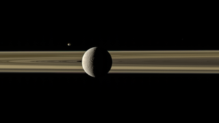 Saturn's moons Rhea and Janus tango on opposite sides of the planet's famous ring system in this new color-enhanced image from NASA's Cassini orbiter. Rhea, Saturn's second-largest moon, is visible in the foreground, while the smaller moon Janus is pictured in the distance across the rings. Citizen scientist Kevin Gill recently processed this 10-year-old view of Saturn and two of its moons using calibrated red, green and blue filtered images captured by the Cassini spacecraft on March 28, 2010.