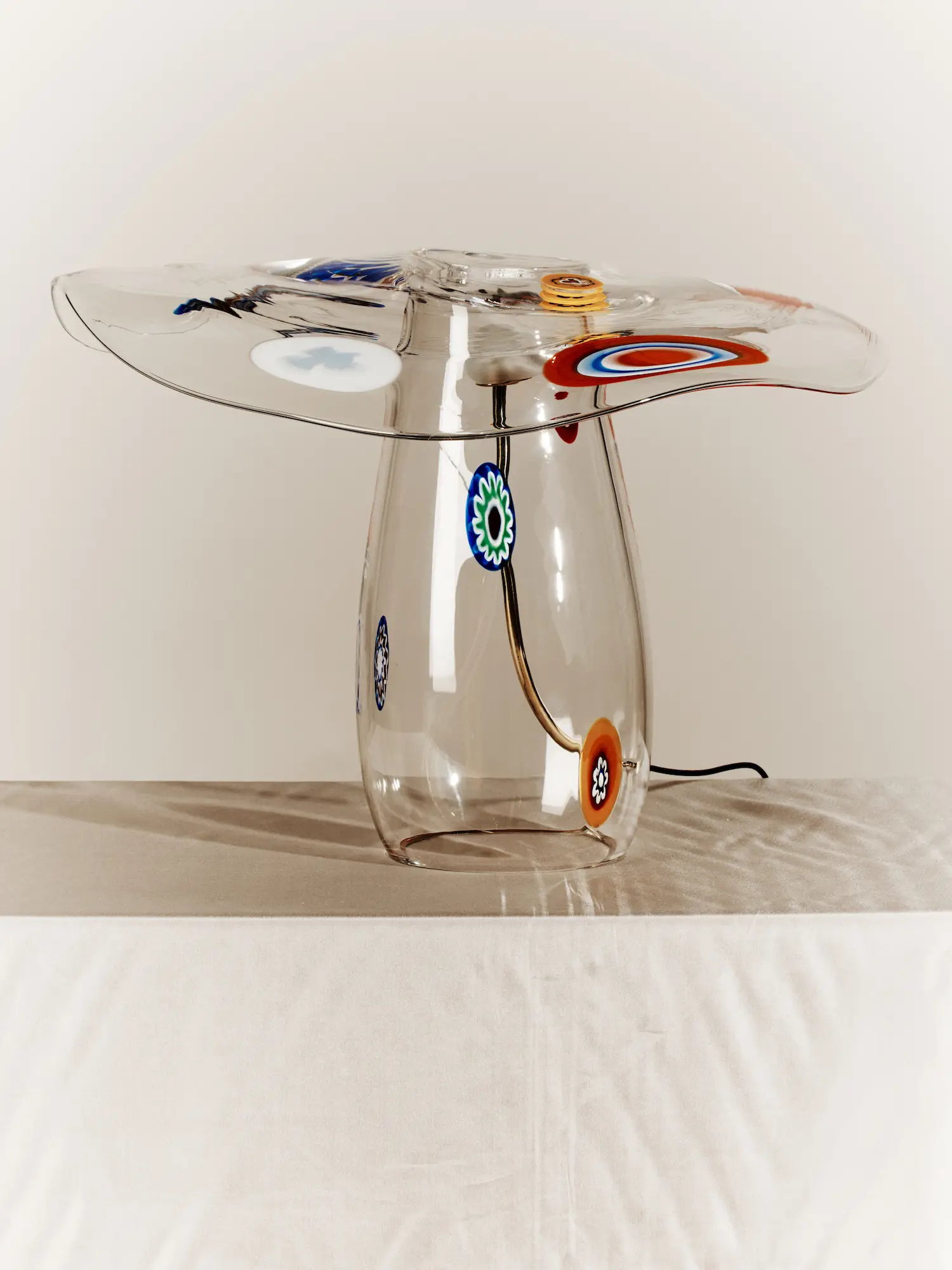 A see-through, mushroom-shaped glass lamp with colorful Venetian 'murine' with floral shapes sits atop a sun-lit table covered in a white cloth in a white room.