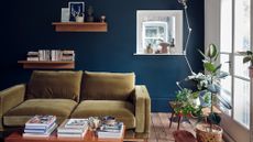 Image of a living room painted in a dark blue. There is an ochre yellow, velvet sofa, a couple of leafy plants by the window on the right, and wooden floating shelves with books on them.