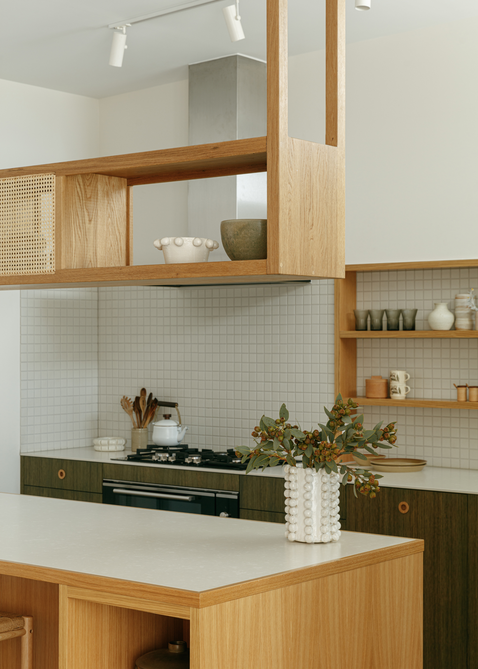A kitchen with a white tile backsplash, wood shelving, green cabinetry, and an island with a bobbin-style vase with stems