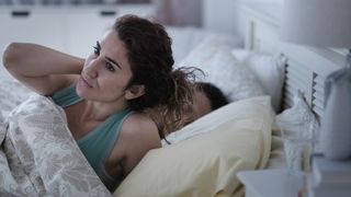 Woman in green bed sits up in bed while her partner sleeps next to her
