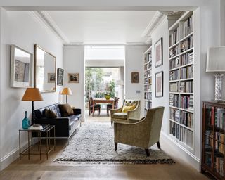 Bright open-plan living and dining space, white painted walls, floor to ceiling shelving filled with books, cozy seating area with sofa and two armchairs, soft gray textured rug underneath, lots of framed pictures and paintings on walls, reading and desk lamps beside seating areas, rectangular wall mirror above sofa