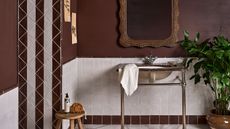 A bathroom with brown walls, white tiles, a brown granite standing sink, a light wooden stall with a white soap dispenser and shower sponge on it, and a patterned white and brown tile strip on the wall