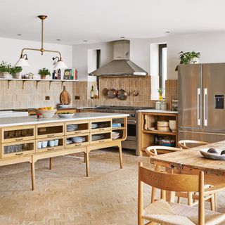 kitchen with terracotta floor tiles, a kitchen island with shelving and drawers, a neutral-tiled kitchen splashback, stainless-steel range cooker and fridge freezer and wooden dining table and chairs