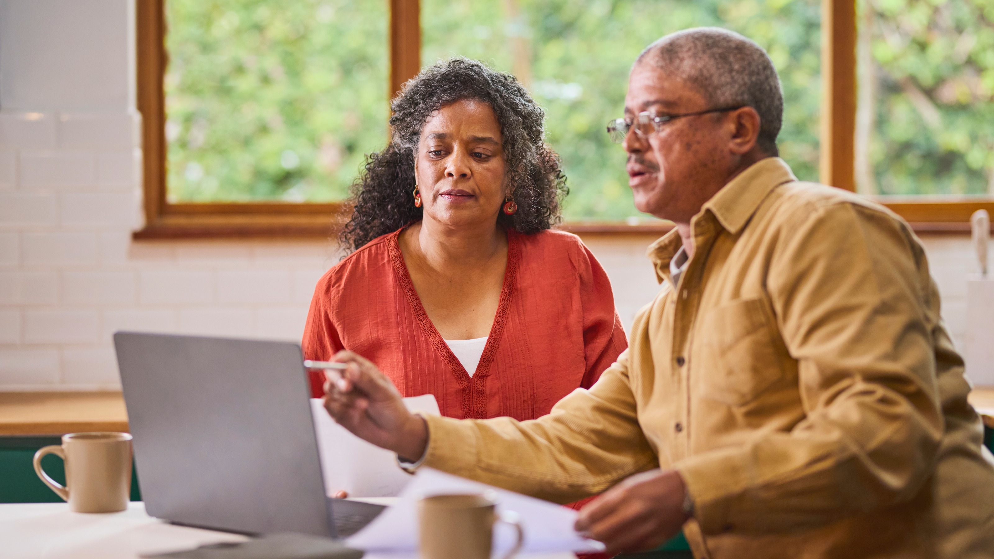 A mature couple works seriously at a kitchen table, reviewing information on a laptop