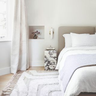 A cream bedroom with a stone column bedside table and a tufted, fringe-trimmed rug