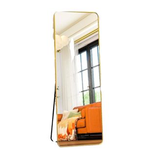 A rectangular full-length mirror with a gold border and black stand at the back, reflecting a living area with white walls, a black full-length window and an orange couch