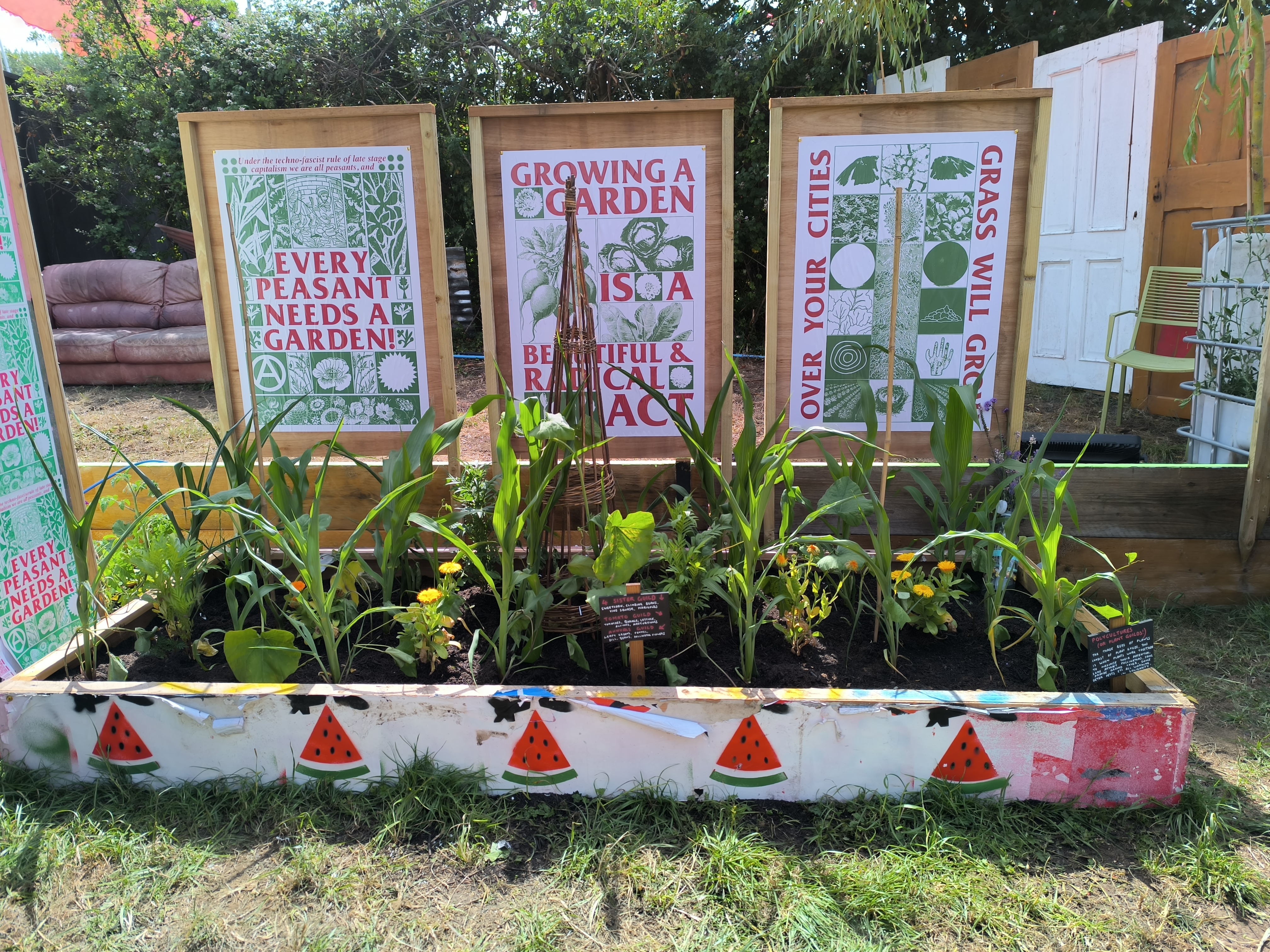 A rectangular wooden garden bed filled with soil and growing plants sits outdoors, featuring a white-painted front border decorated with a row of red watermelon slice graphics.