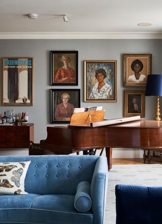 Image of a chic, traditional-style living room. There is a cherry wood grand piano in front of a gallery wall of painted portraits. In the front of the room there is a light blue, velvet sofa with a matching bolster pillow.