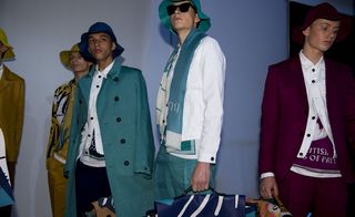 Five male models wearing looks from the Burberry Prorsum collection. One model is wearing a mustard coloured jacket. Another model is wearing mustard coloured trousers and a white, black and mustard jacket. The third model is wearing dark blue trousers, a white and dark blue t-shirt and a white jacket with a blue coat on top. The fourth model is wearing blue trousers, a blue and white t-shirt, a white jacket and a dual coloured scarf. The fifth model is wearing a white and maroon t-shirt, white jacket and maroon suit. All models are wearing hats, one is wearing sunglasses and some are holding briefcases