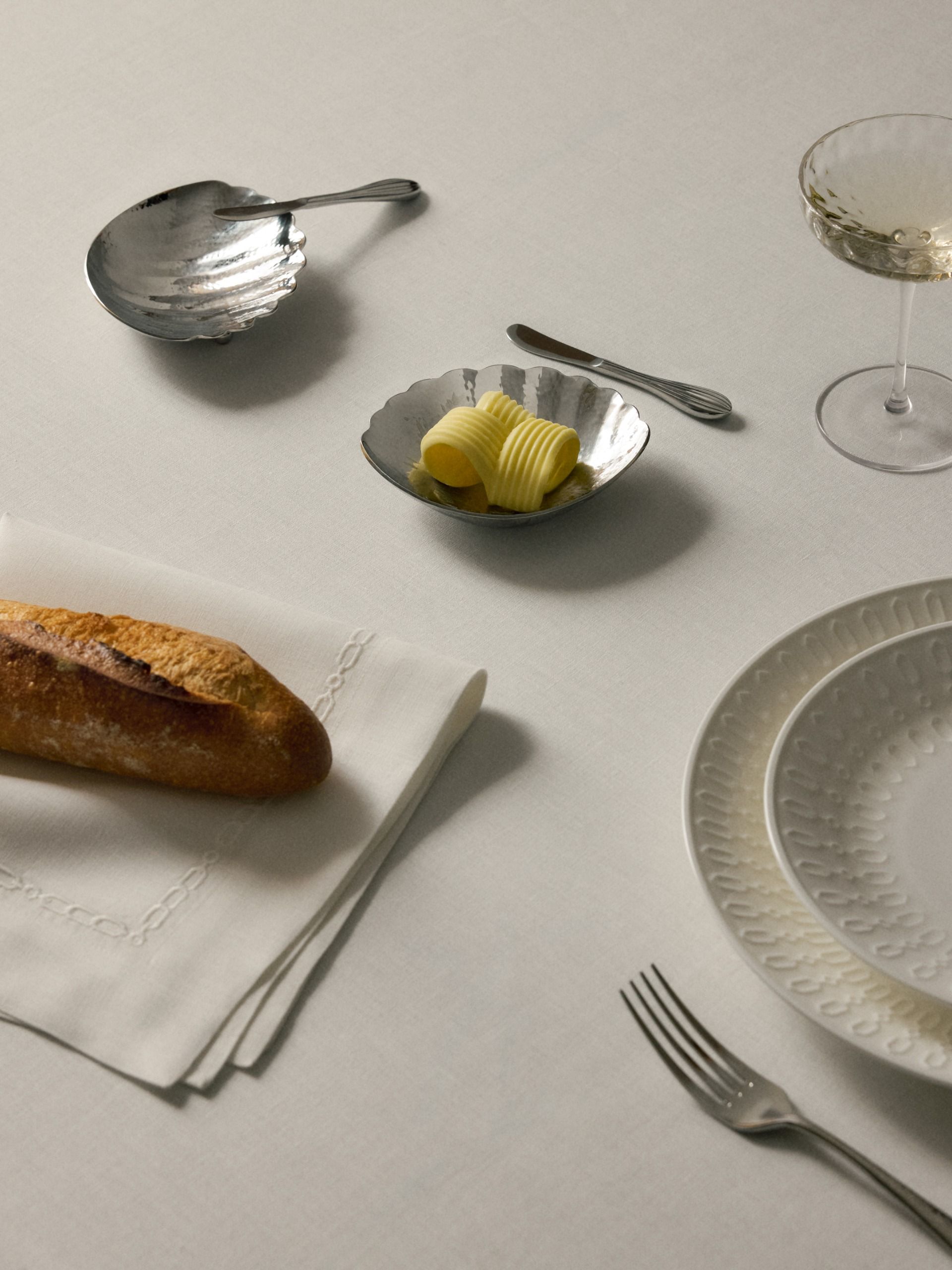 close up of table with white tablecloth, chrome shell-shaped butter holder, embossed dinnerware, silverware, napkin and baguette