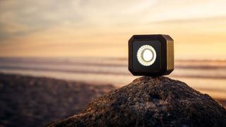 Lume Cube Air, a small light cube, shown on a small hill with a beach at sunset behind