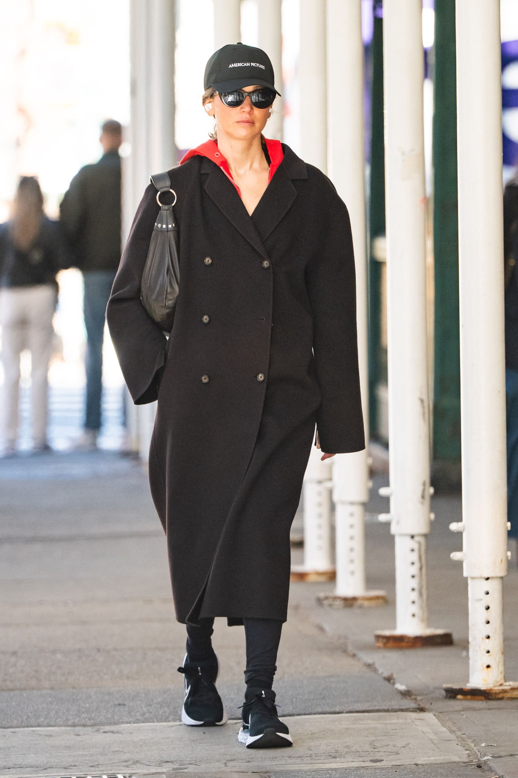 Jennifer Lawrence wears a baseball cap, brown wool coat, red sweatshirt, black leggings, and black sneakers.