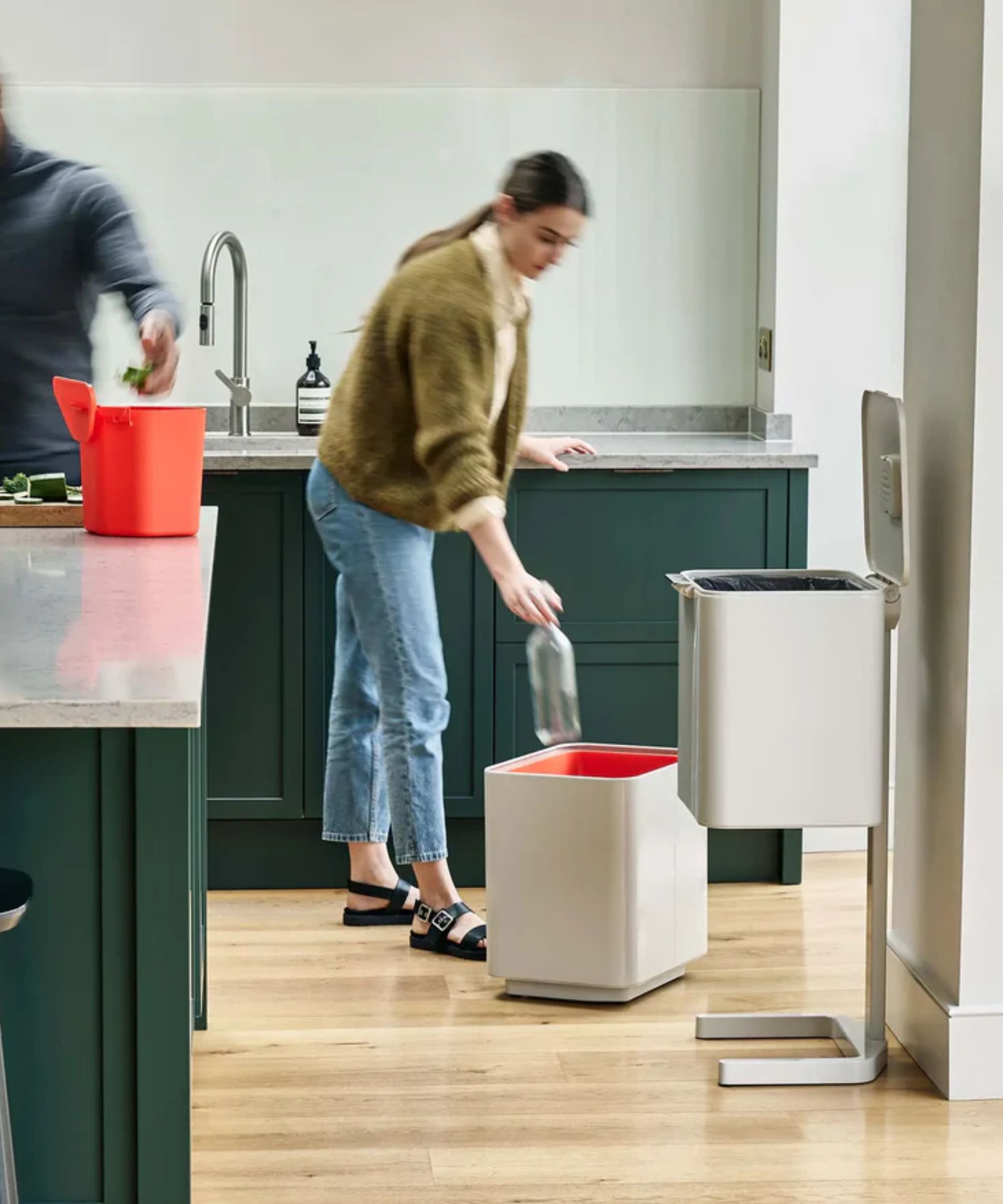 The white version of the Joseph Joseph Totem Trash Can in a kitchen, separated into its three parts and being used by two people. One throwing an empty bottle into the bottom compartment, and one putting food waste in the removeable compost bin.