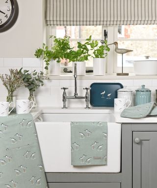 A coastal kitchen with a large window with striped blinds and plants, a bird figure, and a white pot on the windowswill, and a white sink unit with mint tea towels on it and gray cabinets