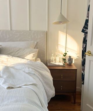 A white bedroom with wall paneling, a bed with white sheets and pillows, a dark brown nightstand with bottles on top of it, and a white pendant light over it