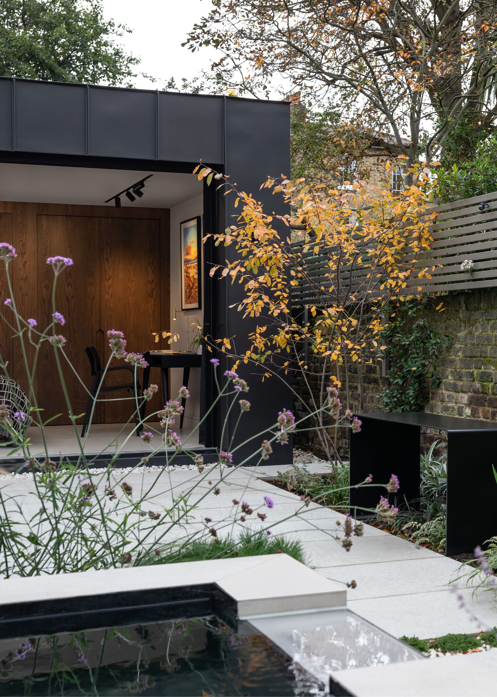 An area of the patio in a home with a view of lots of plants alongside borders. There is also a black bench on the right side of the border by the brick walls. You can also see a glimpse of the garden room that has a desk and chair inside. 