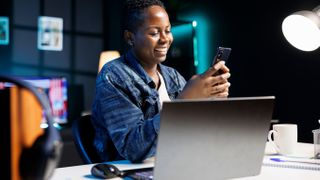 A woman using a phone while she's sitting at a desk that has a laptop on it