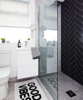 A modern monochrome black and white bathroom with shower, Herringbone tiling, waterfall shower and 'Good Vibes' statement bath mat