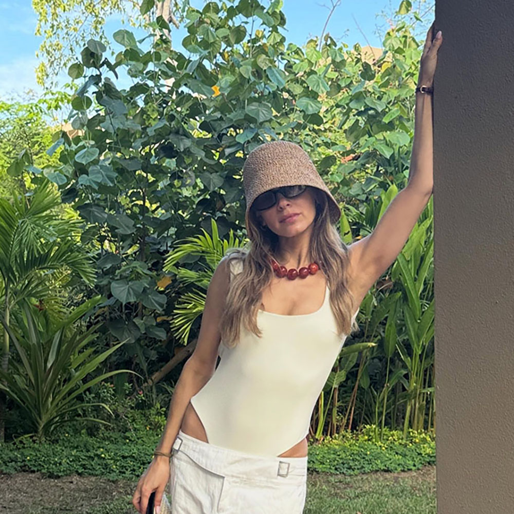 a woman wearing a woven bucket hat, red beaded necklace, white one-piece swimsuit, and white pants