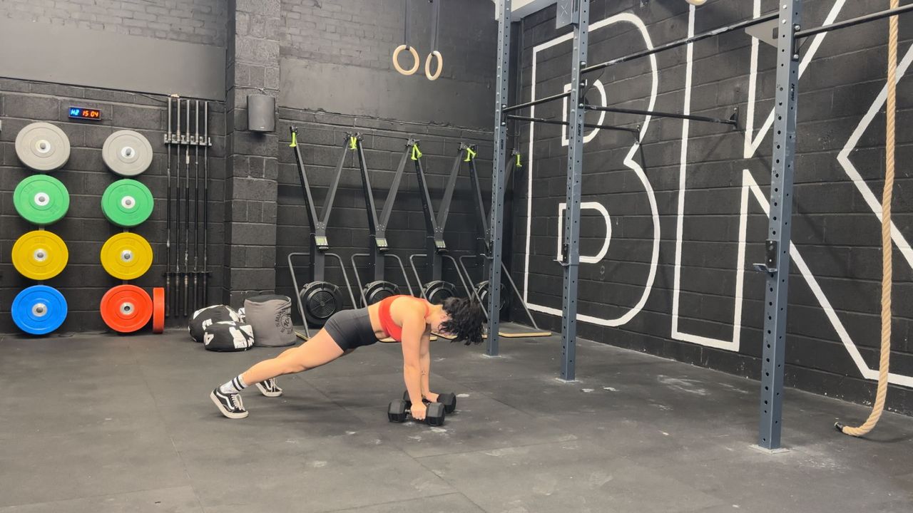 Raquel Sanjurjo performs a renegade row into triceps kickback in a gym. She is in a high plank position holding a pair of dumbbells on the ground.