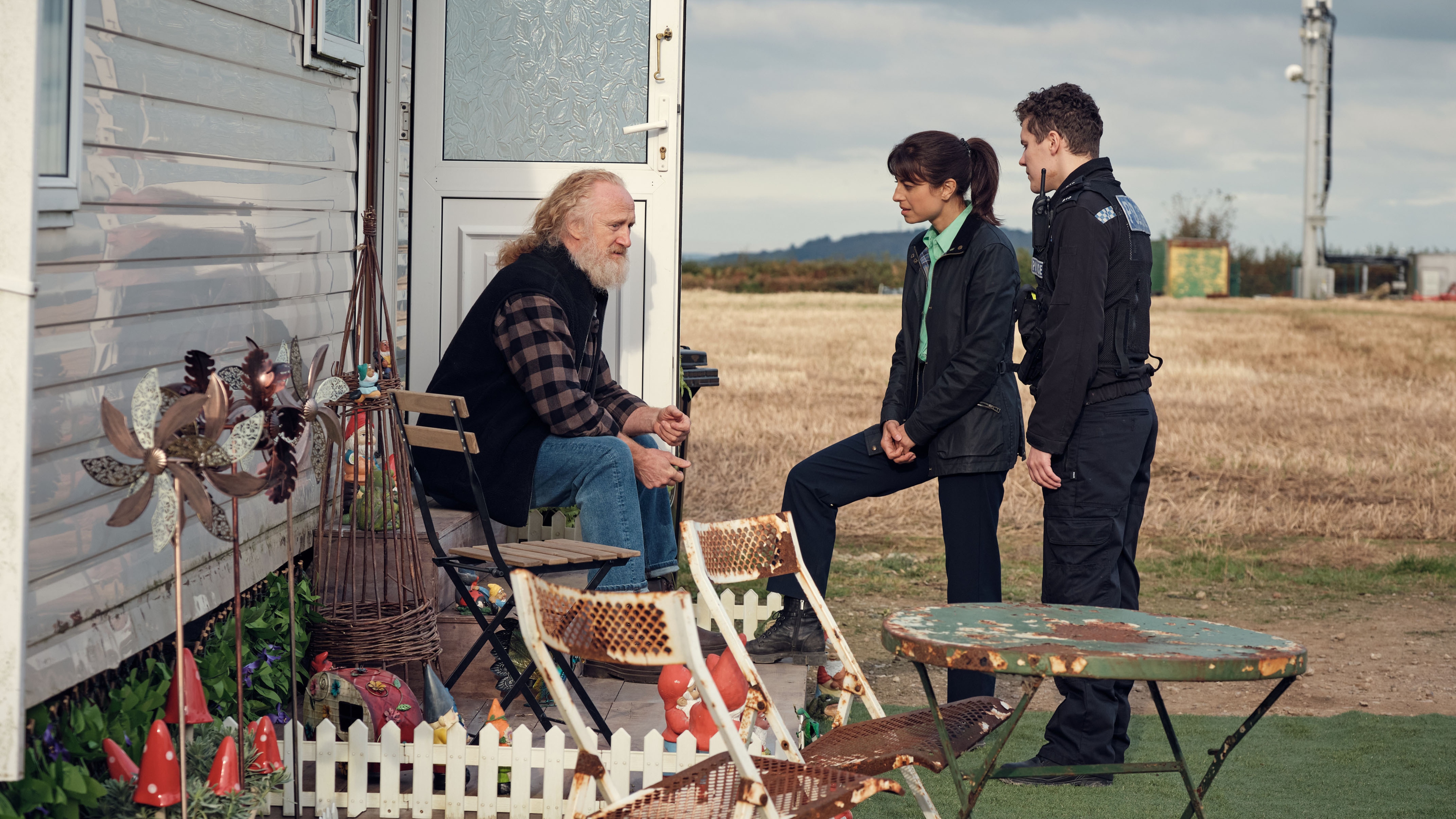 Trent Morgan (Murray McArthur) sits on the steps of his static caravan with the door open, while DS Esther Williams (Zahra Ahmadi) and PC Kelby Hartford (Dylan Llewellyn) stand facing him. There are some rusty-looking garden chairs and a table in the front yard and a small garden area with a mini white picket fence surrounding it.
