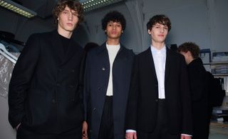Four male models wearing looks from Margaret Howell's collection. Two models are wearing black suits with one wearing a black top and the other wearing a white shirt. Another model is wearing a white top, black trousers and blue coat. The fourth model is behind the main three and is wearing a black coat and backpack