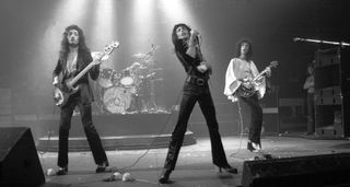 Queen live in 1974, four white stage lights illuminating Roger Taylor's drum kit as Freddie Mercury fronts the band, flanked by John Deacon on the left and Brian May on the right.