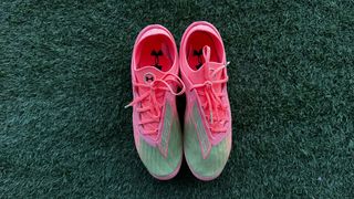 A bird's eye view of pink and green football boots with pink laces and the Under Armour logo on the sole, sitting on green artificial grass. Under Armour Clone Magnetico Elite 4.0