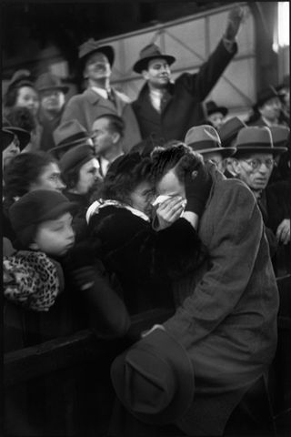 Black and white image of New York City,1946. Close up shot of a crowd of people in smart dress, some men in dark colour trilby hats, older women hugging a younger man who is on the other side of a partition fence, holding a tissue to his face