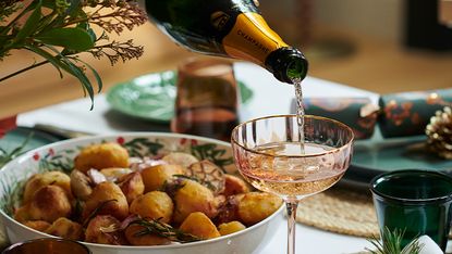 A glass of champagne being poured into a glass with a bowl of roast potatoes in the background.