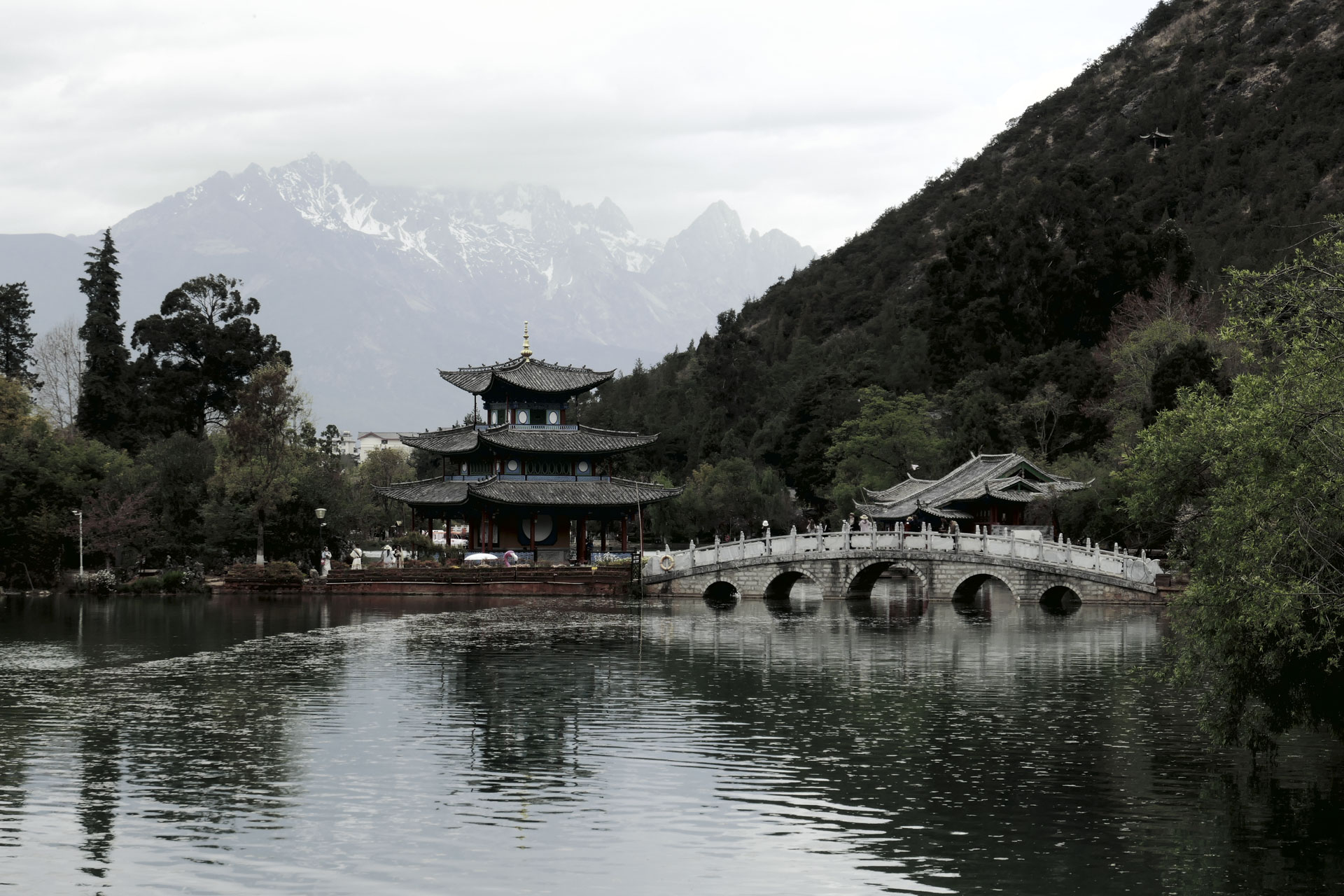 A Chinese temple by the side of a lake with an arched bridge leading up to it and both are reflected in the still water, in front of a snow covered mountain. A creative filter is applied to the image