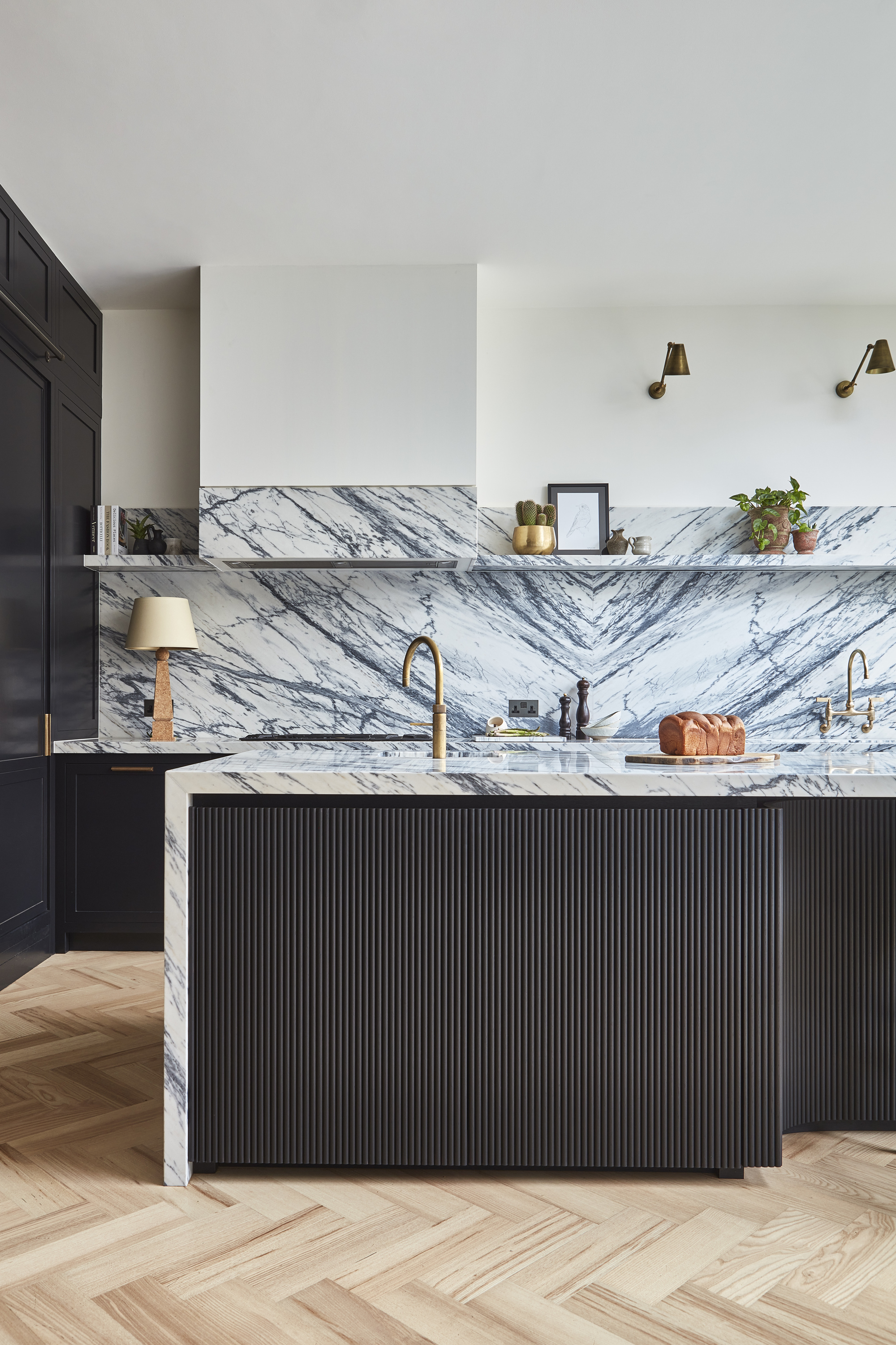A kitchen with a bookmatched marble countertop, wooden herringbone floors, and a black fluted cabinetry