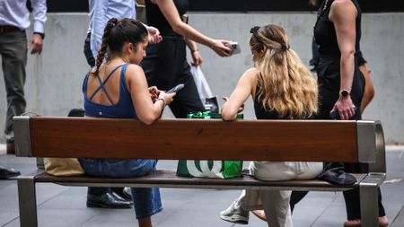 Pedestrians walk past two young women sitting on a bench looking at their phone in central Sydney 