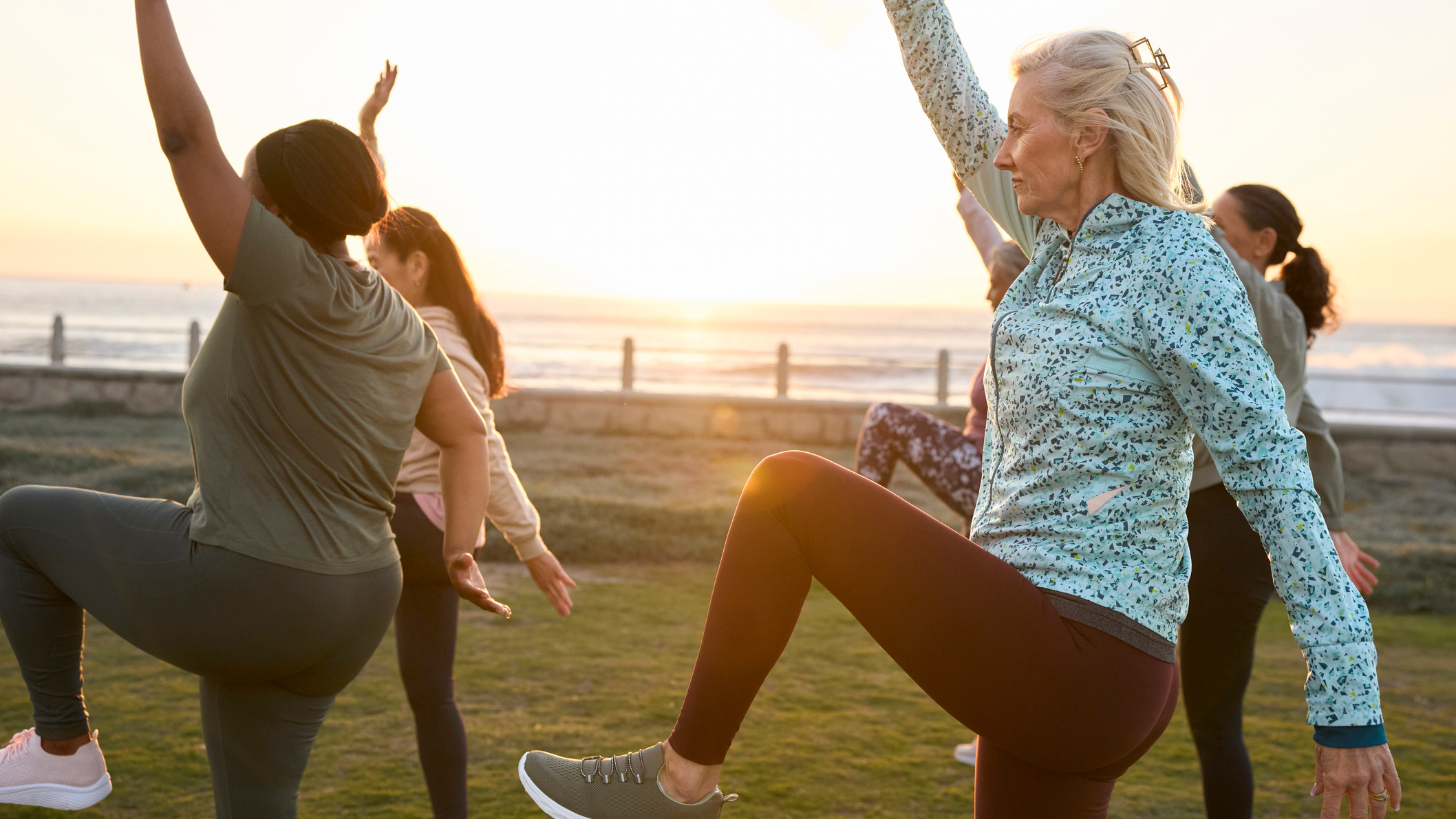 A group of women exercising outdoors, with their left legs and right arms raised