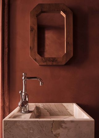 Image of a marble bathroom sink. The walls are painted in a black-orange paint color using a limewashing technique. There is a wooden statement mirror hanging over the sink.