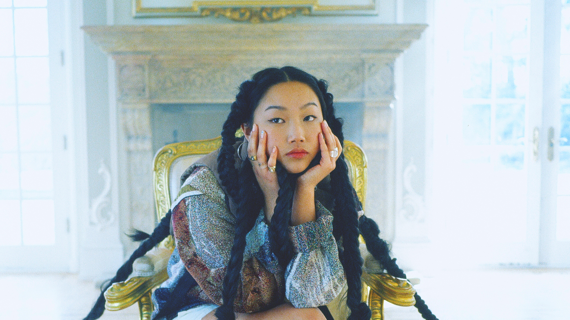Musician Audrey Nuna sitting in an ornate, gold-accented armchair with her chin resting on both hands, wearing long braided pigtails in front of a fireplace.