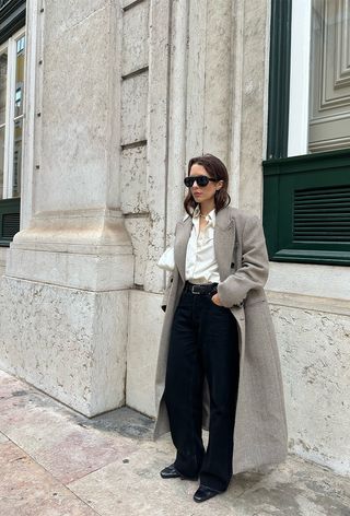 A woman standing outside wearing a long tan coat over a white button-down shirt with black jeans, black block low-heel boots, a black belt, and a white shoulder bag.