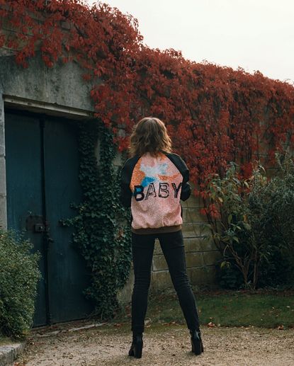 A female model standing in a garden, back turned to the camera, wearing a a top with block sleeves and a colourful body with the word "Baby" written across the back in a stencil font.