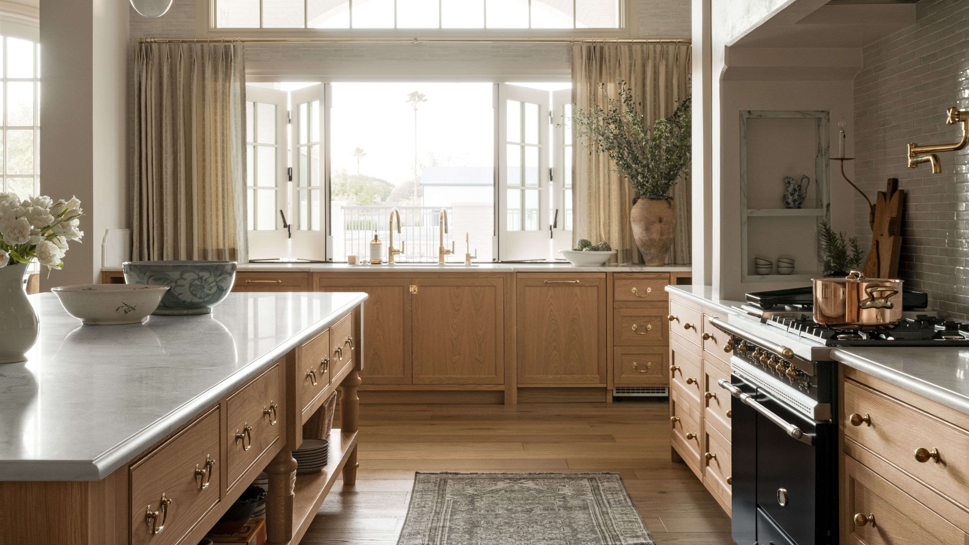 a large open oak kitchen with a large open window, drapes, a sink at the window with a large vase of foliage. to the left is a huge island and to the right a cooker with built in hood and storage