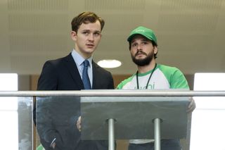 Robert (Harry Lawtey) and Henry (Kit Harington) stand on a balcony at the Stock Exchange with a terminal in front of them. Henry is wearing a green Lumi-branded cap and t-shirt.