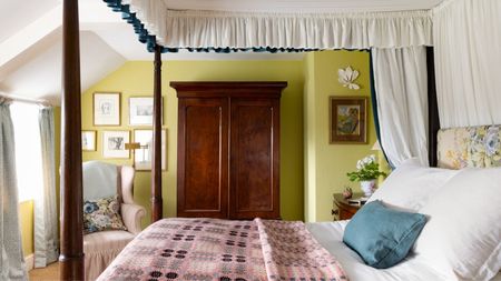 Bedroom with citron green painted walls, a mahogany four-poster bed with a canopy, a floral headboard and a pink Welsh blanket, and a large mahogany wardrobe beside a pale pink armchair.