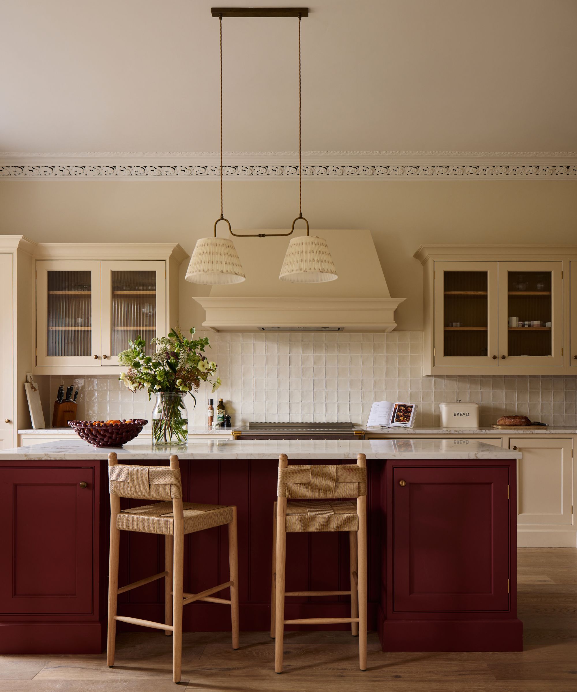 A deep red kitchen island with two woven wooden barstools under a double fabric pendant light, backed by cream cabinets and a stove range