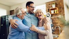 Older parents happily hug their adult son in the living room.