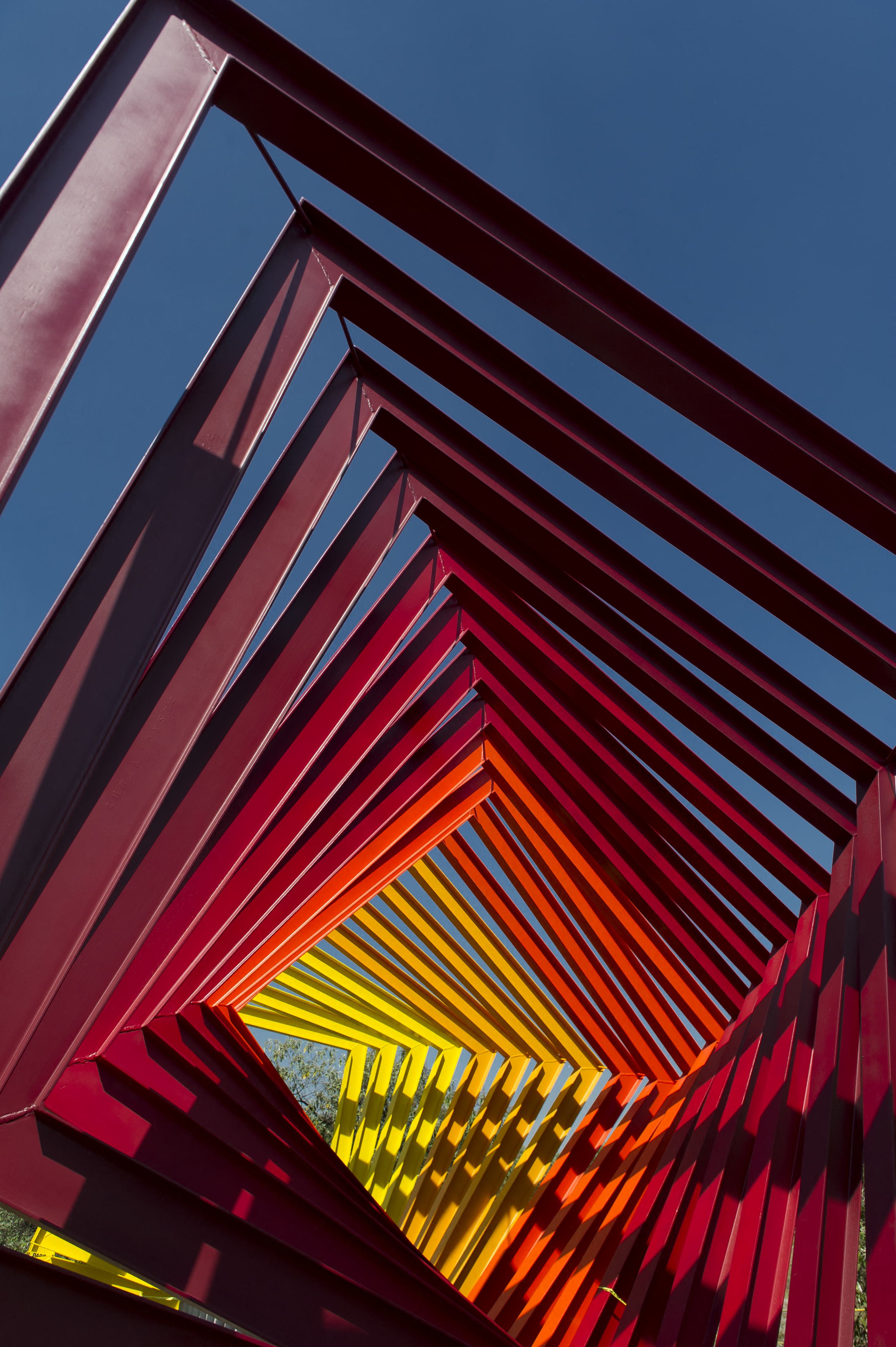 A colorful, red, orange, and yellow spiraling sculpture sits in a park in the sun.